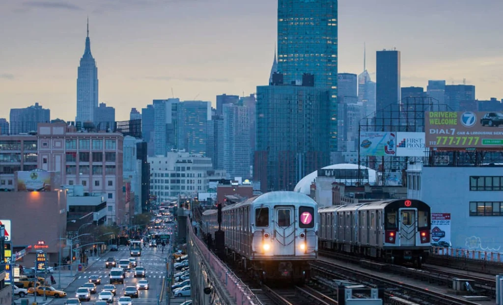 Twee metrotreinen rijden over een verhoogd spoor bij avondlicht met het Empire State Building op de achtergrond