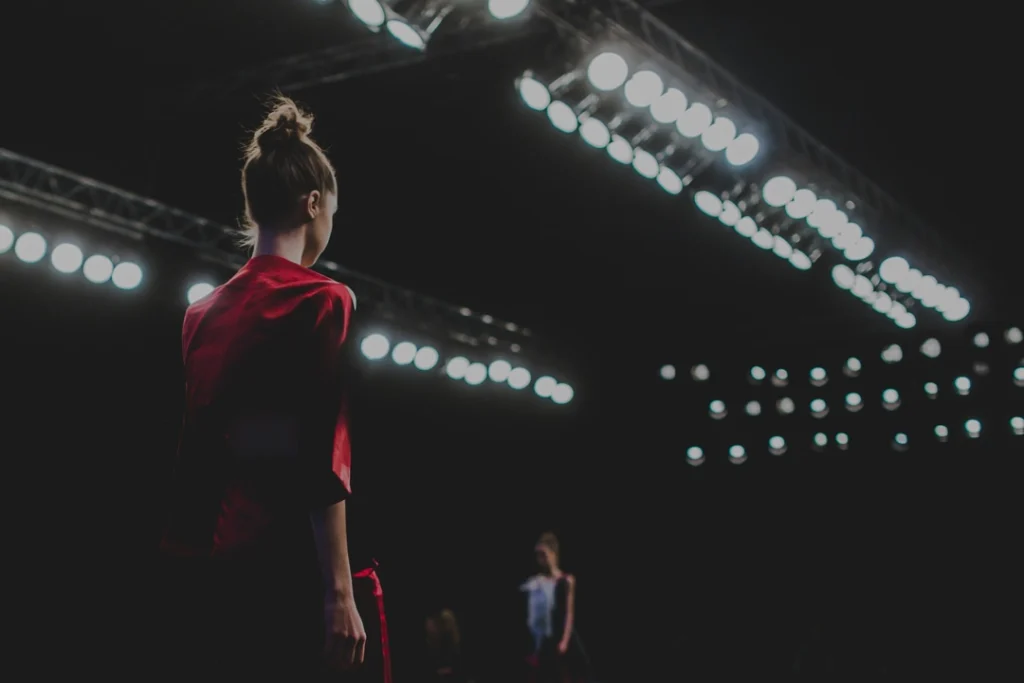Model met opgestoken haar in een rode outfit loopt van achteren gezien onder felle spotlights over de catwalk