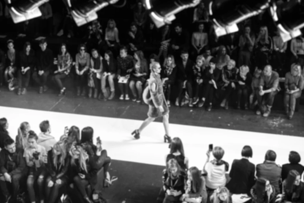 Zwart wit foto van een model dat over een verlichte catwalk loopt tussen zittend publiek