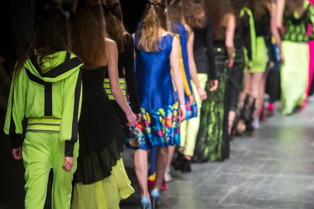 Rij modellen loopt van achteren gezien in felgele groene en blauwe outfits over de catwalk