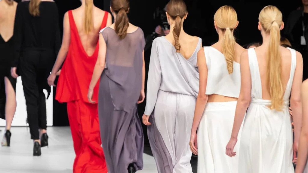 Rij modellen met opgestoken haar loopt van achteren gezien in lange jurken in rood grijs en wit over de catwalk
