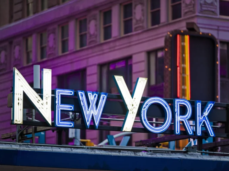 Blauw en wit neonbord met de tekst New York bevestigd aan een lantaarnpaal voor een paars verlicht gebouw bij schemering