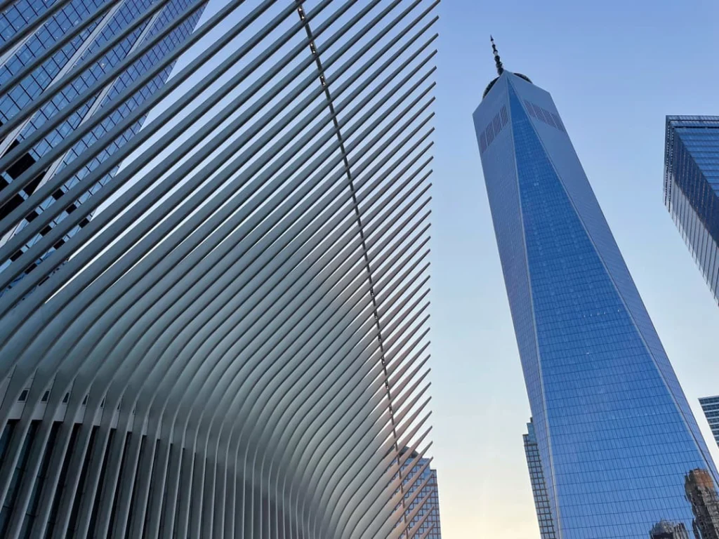 Van onderaf gefotografeerde witte ribbenstructuur van de Oculus naast de glazen torenspits van One World Trade Center
