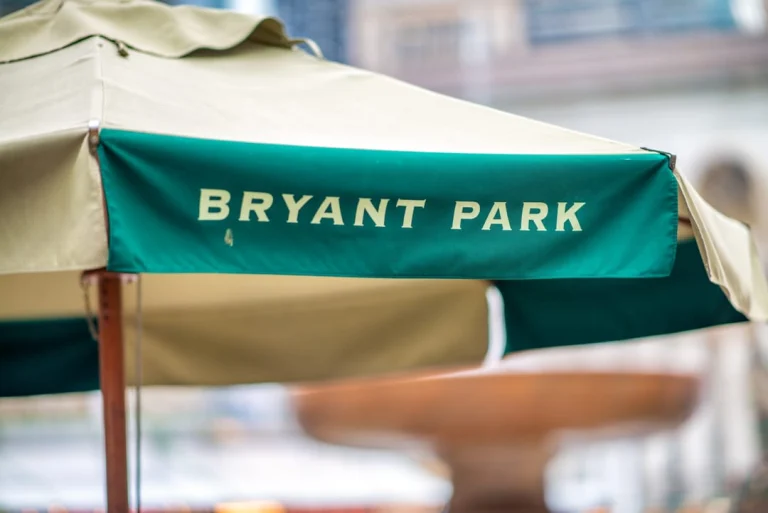 Close up van een groene en beige parasol met daarop de tekst Bryant Park boven een rond houten tafeltje