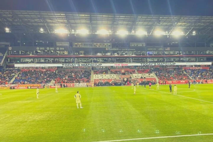 Voetballers warmen op op het veld van Red Bull Arena terwijl het publiek op de tribunes de wedstrijd volgt