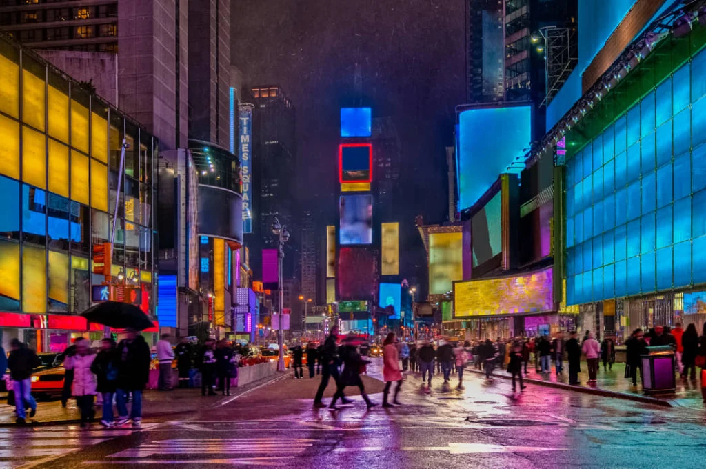 Nat wegdek weerkaatst het felle neonlicht van reclameborden op Times Square, terwijl voetgangers met paraplu's oversteken in de regen