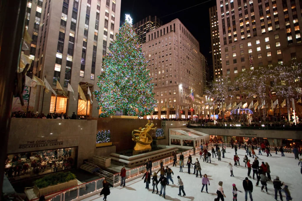 Drukke ijsbaan met schaatsers in de avond, omringd door hoge gebouwen en een grote verlichte kerstboom bij het Rockefeller Center