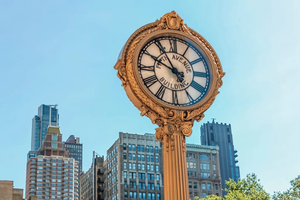 Gouden antieke straatklok met de tekst Fifth Avenue Building tegen een blauwe lucht met wolkenkrabbers op de achtergrond
