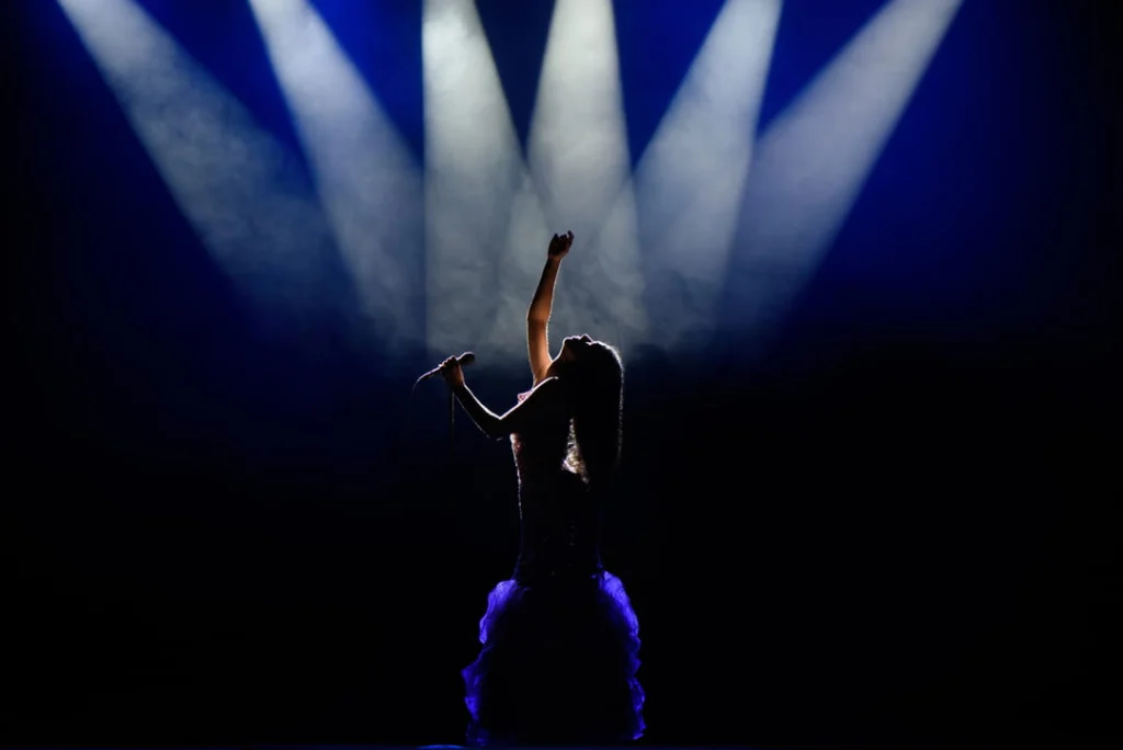Silhouet van een vrouw in jurk die met geheven arm zingt terwijl blauwe spotlights door rook schijnen op een podium