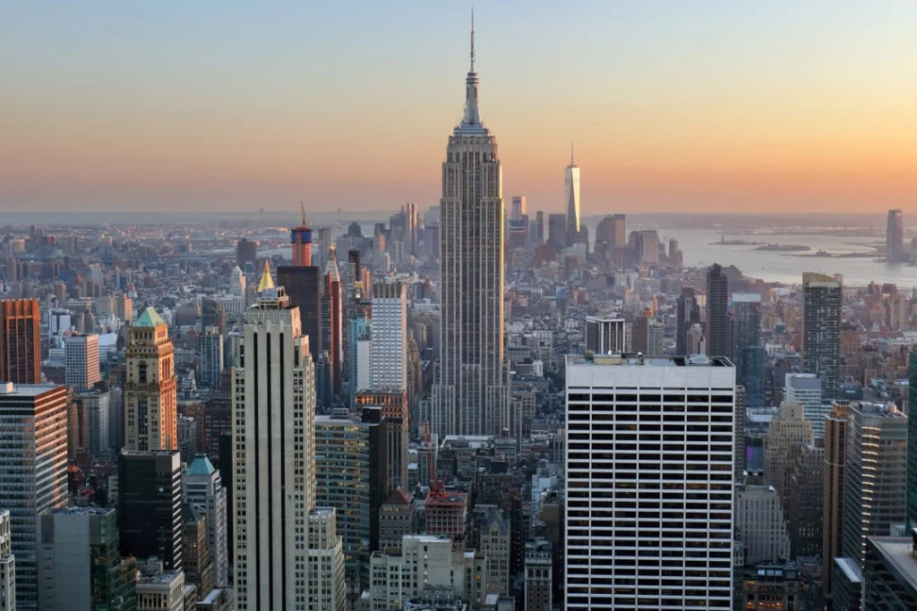 Uitgestrekte skyline van Manhattan bij zonsondergang met het Empire State Building en One World Trade Center en water van de haven op de achtergrond