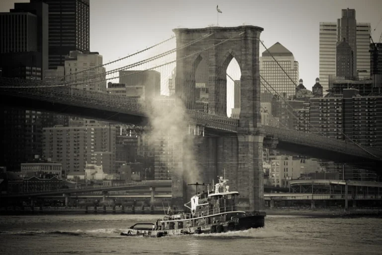 Zwart wit foto van een stomende sleepboot die onder de Brooklyn Bridge vaart met de skyline van Manhattan op de achtergrond