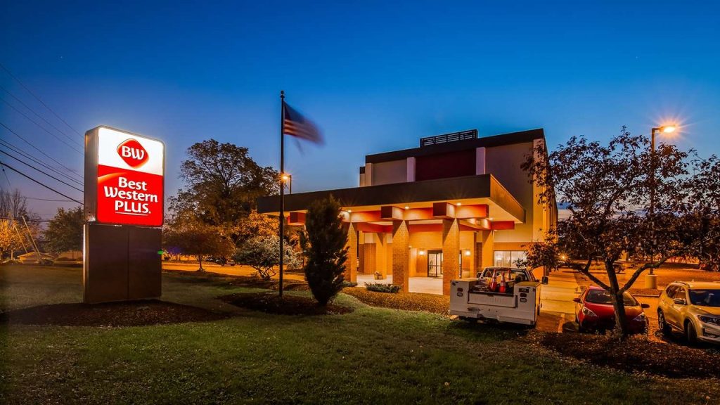 Gevel van een Best Western Plus hotel met verlichte luifel, vlaggenmast en geparkeerde auto's bij avondschemering