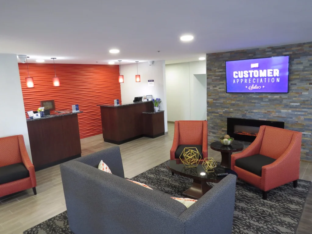 Lobby van een hotel met een oranje gestructureerde wand, een tv, een openhaard en rode en grijze zitfauteuils