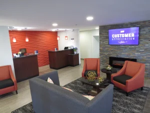 Lobby van een hotel met een oranje gestructureerde wand, een tv, een openhaard en rode en grijze zitfauteuils