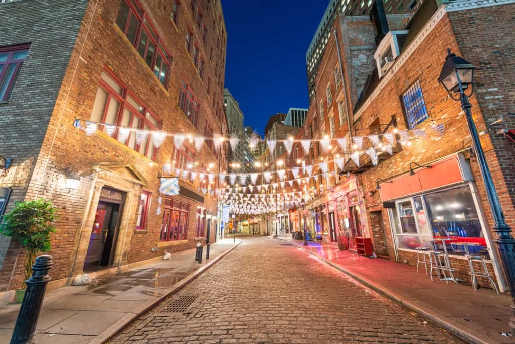 Kasseienstraat Stone Street in New York met rijen witte vlaggetjes tussen bakstenen gebouwen en wolkenkrabbers op de achtergrond bij nacht