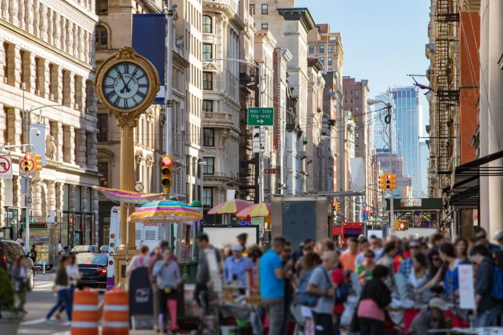Levendig straatbeeld op Fifth Avenue met een gouden sierklok, marktkraampjes en veel voetgangers tussen historische gebouwen