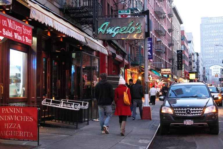 Voetgangers lopen over de stoep langs restaurants en winkels met verlichte uithangborden in Little Italy in New York
