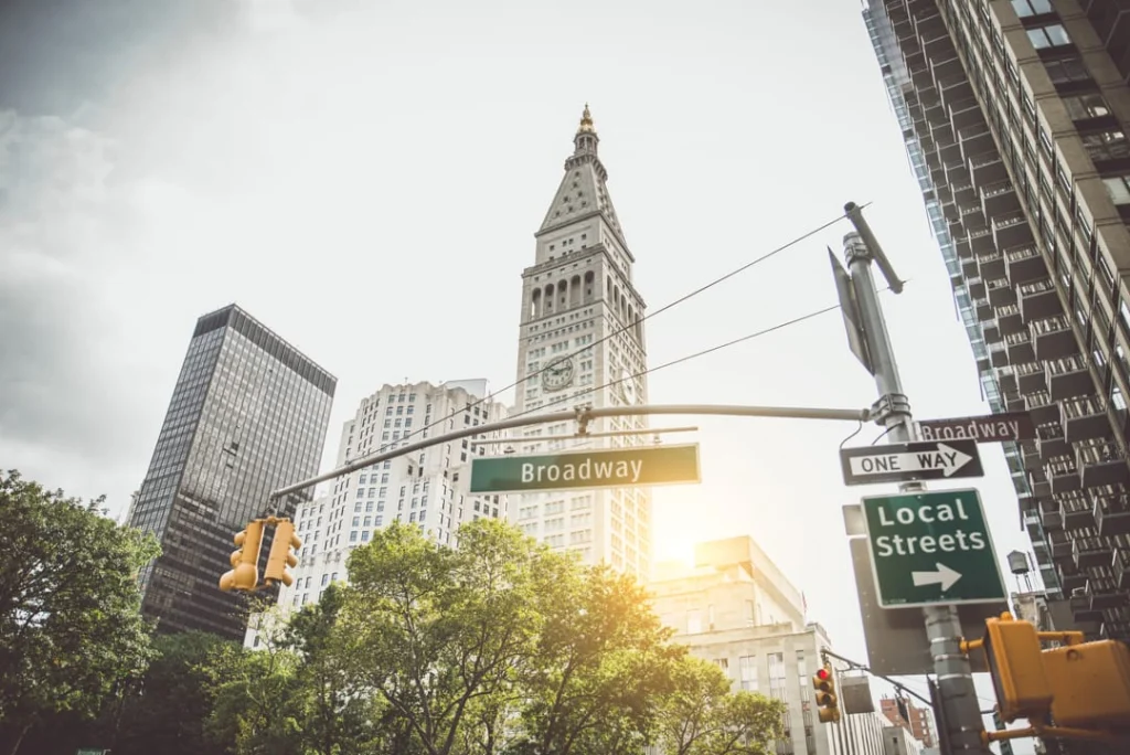 Groen straatbord met de tekst Broadway aan een verkeerslicht met een hoge klokkentoren en wolkenkrabbers op de achtergrond