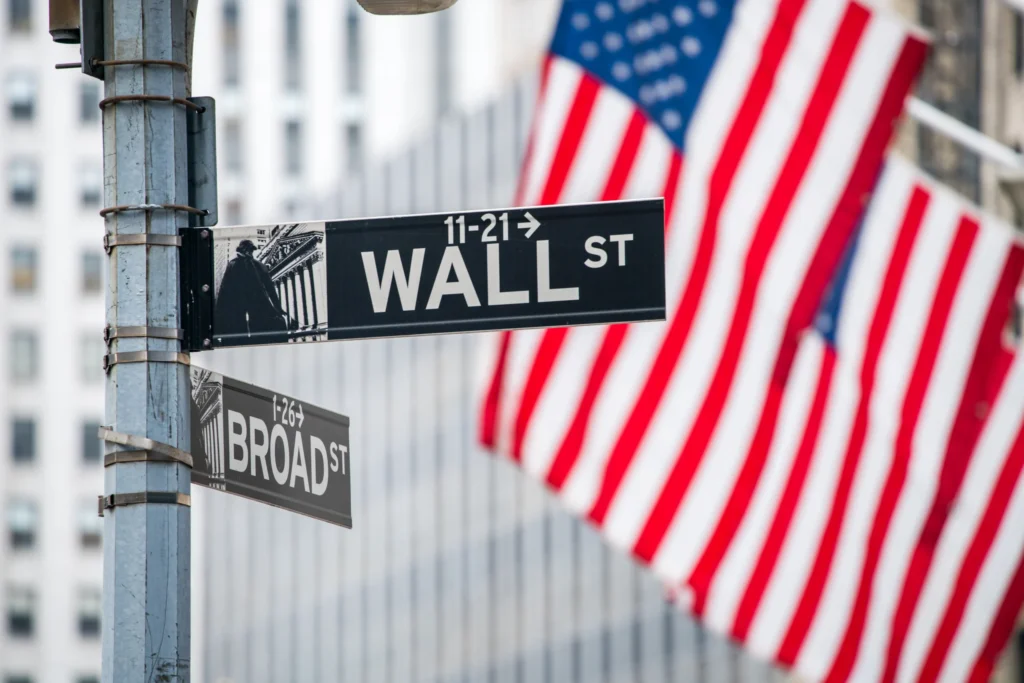 Zwart straatnaambord met de tekst Wall Street aan een lantaarnpaal met wapperende Amerikaanse vlaggen op de achtergrond