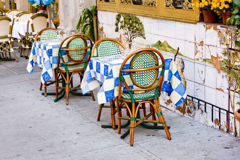 Terrastafels met blauw wit geblokte kleedjes en rieten stoelen voor een beschilderde gevel in Little Italy