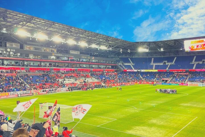 Volle tribune van de Red Bull Arena met zwaaiende supportersvlaggen en het speelveld op de achtergrond onder een blauwe lucht