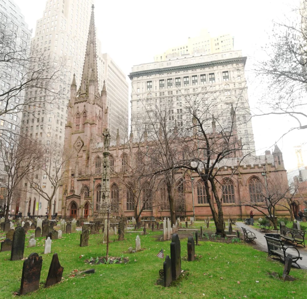 Oude grafstenen op de begraafplaats van Trinity Church tussen kale bomen, met de gotische kerk en kantoorgebouwen op de achtergrond