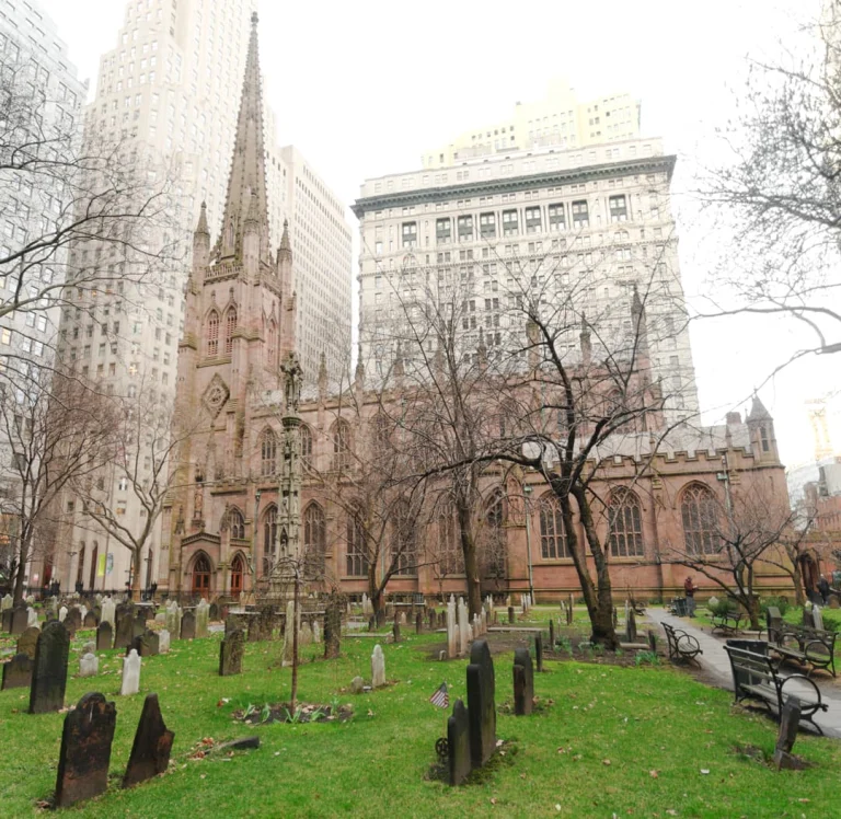 Oude grafstenen op de begraafplaats van Trinity Church tussen kale bomen, met de gotische kerk en kantoorgebouwen op de achtergrond