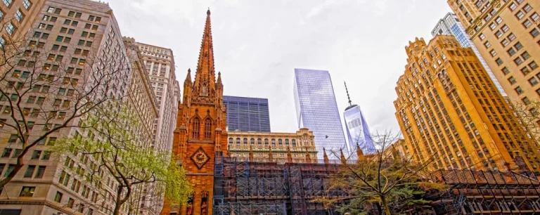 Gotische kerktoren van rode zandsteen met een puntige spits, geflankeerd door hoge kantoorgebouwen van Lower Manhattan onder een bewolkte hemel