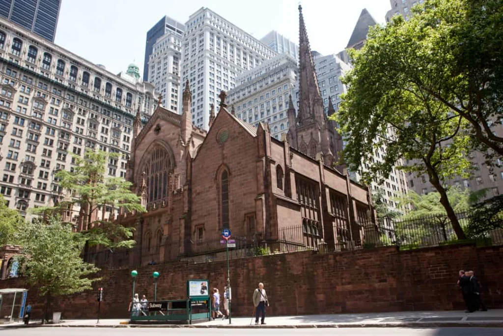 Trinity Church met rode zandstenen gevel aan Broadway in Lower Manhattan New York, met een metro ingang op de stoep ervoor en wolkenkrabbers op de achtergrond