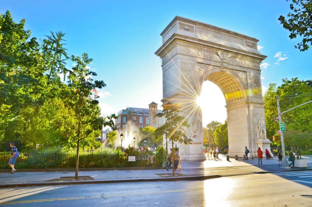 Witte stenen triomfboog van Washington Square Park met de zon die er fel doorheen schijnt en voetgangers op straat