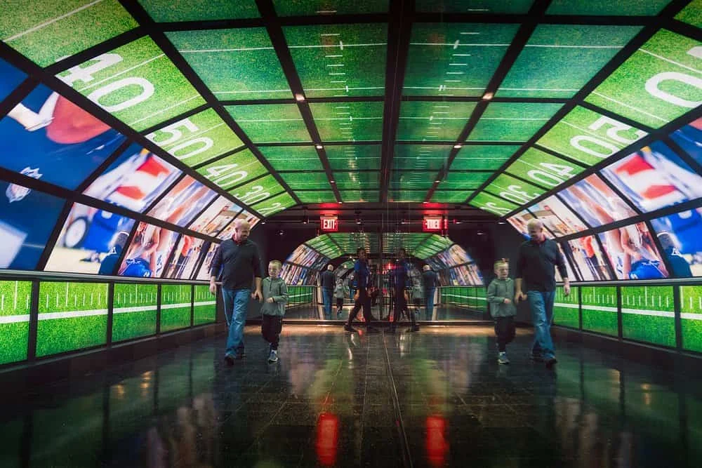 Bezoekers wandelen door een futuristische tunnel met grote schermen die een americanfootballveld tonen in New York