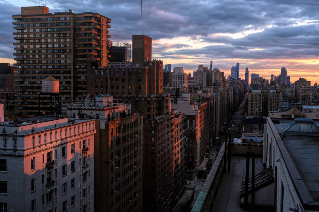 Uitzicht vanaf een dak over torenflats en rijtjeshuizen van New York onder een kleurrijke bewolkte lucht bij zonsondergang