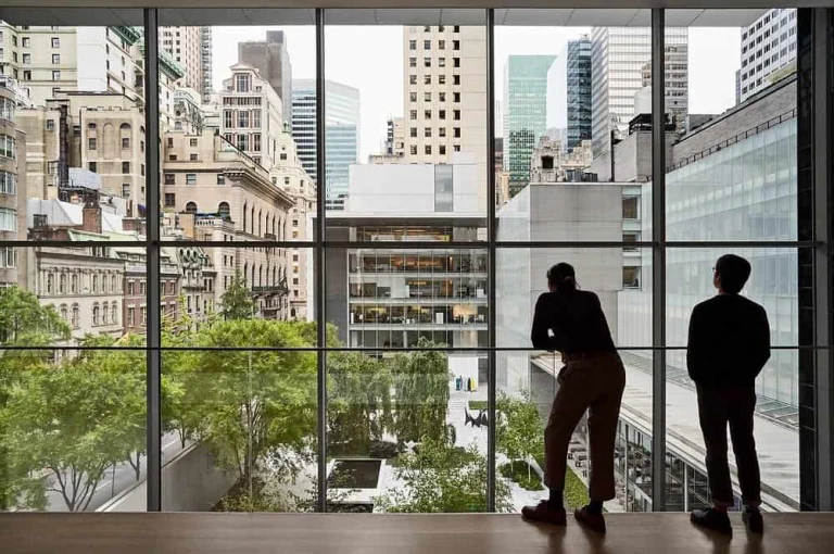 Twee bezoekers kijken door een groot raam van het Museum of Modern Art in New York naar de wolkenkrabbers van Manhattan
