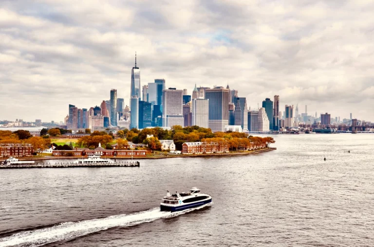 Een veerboot vaart over de rivier voor de skyline van Lower Manhattan met een klein eiland met herfstbomen op de voorgrond