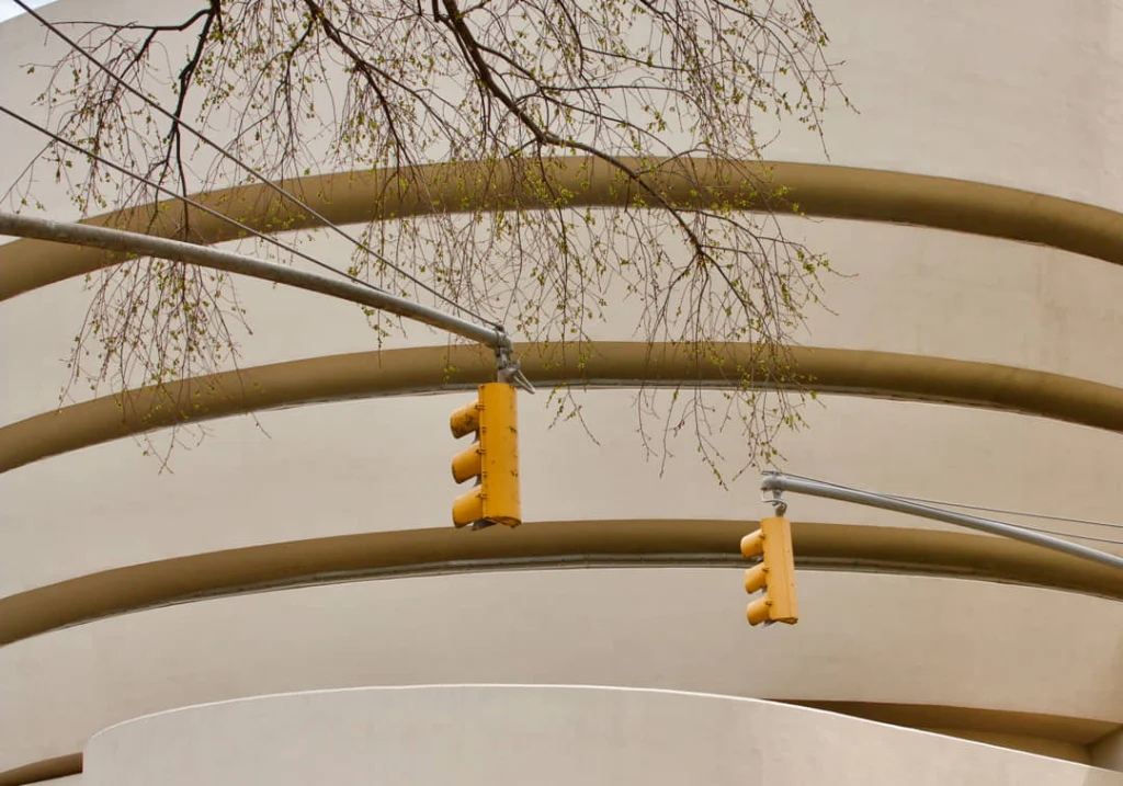 Twee gele voetgangersverkeerslichten aan een lantaarnpaal met boomtakken en de gebogen gevel van het Guggenheim Museum op de achtergrond