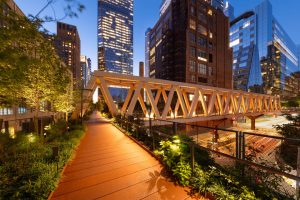 Houten wandelpad met een verlichte stalen brugconstructie op de High Line tussen wolkenkrabbers in de avond in New York