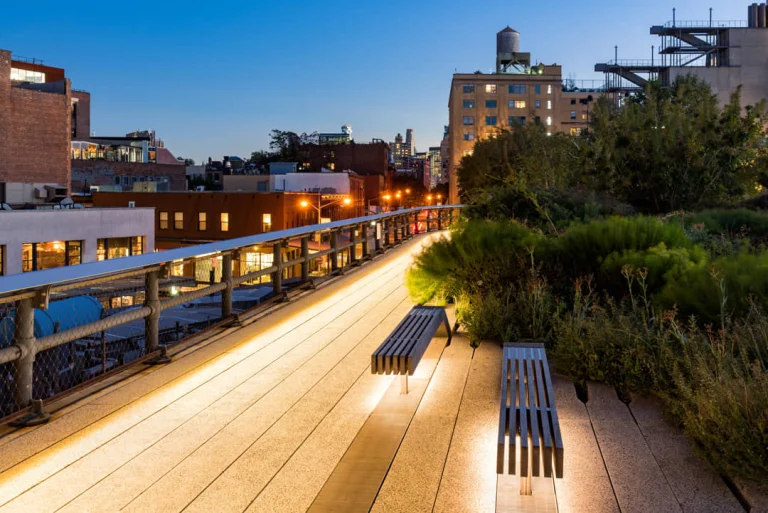 Houten bankjes met ingebouwde verlichting langs het pad van de High Line in de schemering met gebouwen en een watertoren op de achtergrond