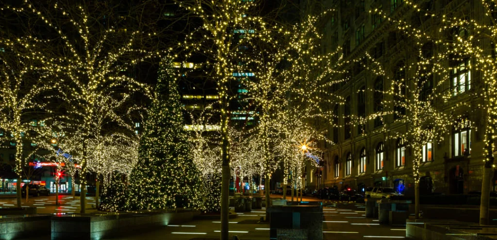 Bomen vol witte lichtjes op een verlicht plein in New York met gebouwen op de achtergrond in de avond