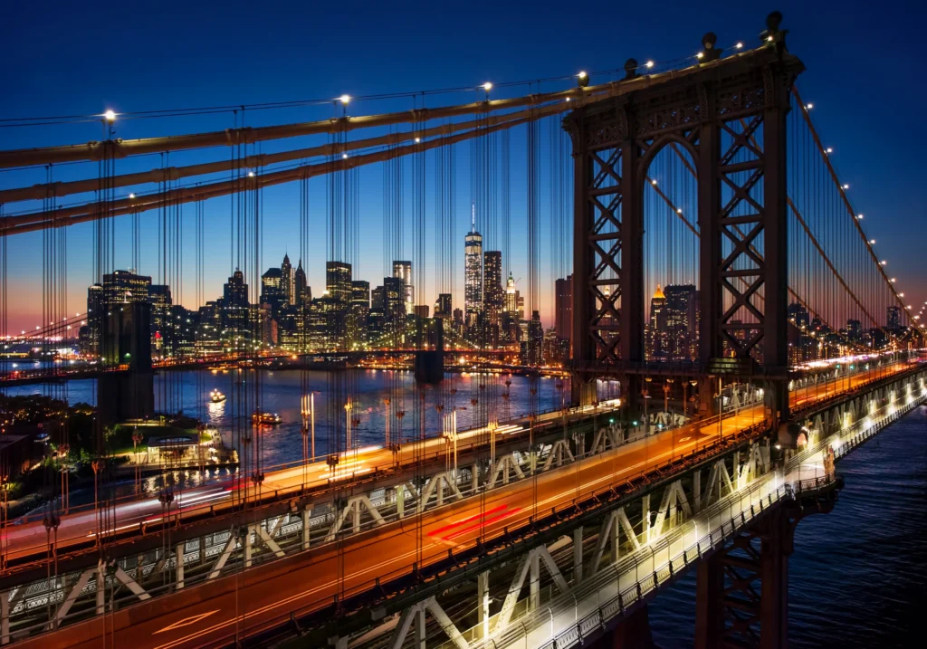 Verlichte hangbrug in New York in de avondschemering met wazige lichtsporen van het verkeer en de skyline op de achtergrond