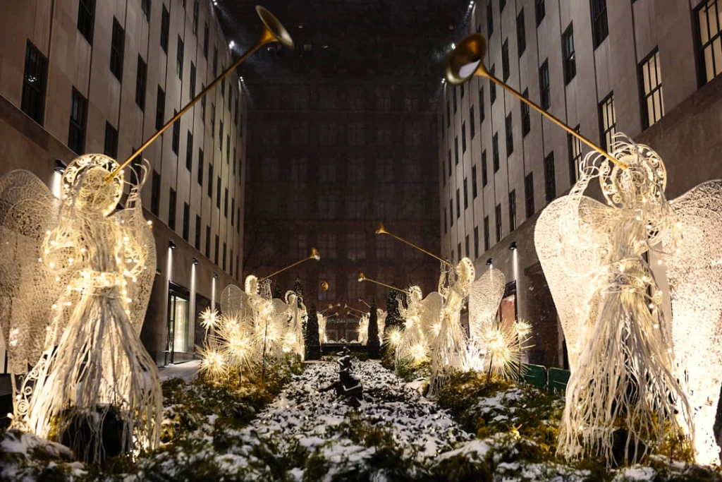 Rij verlichte engelenbeelden met trompetten langs een besneeuwd pad tussen gebouwen bij Rockefeller Center in New York