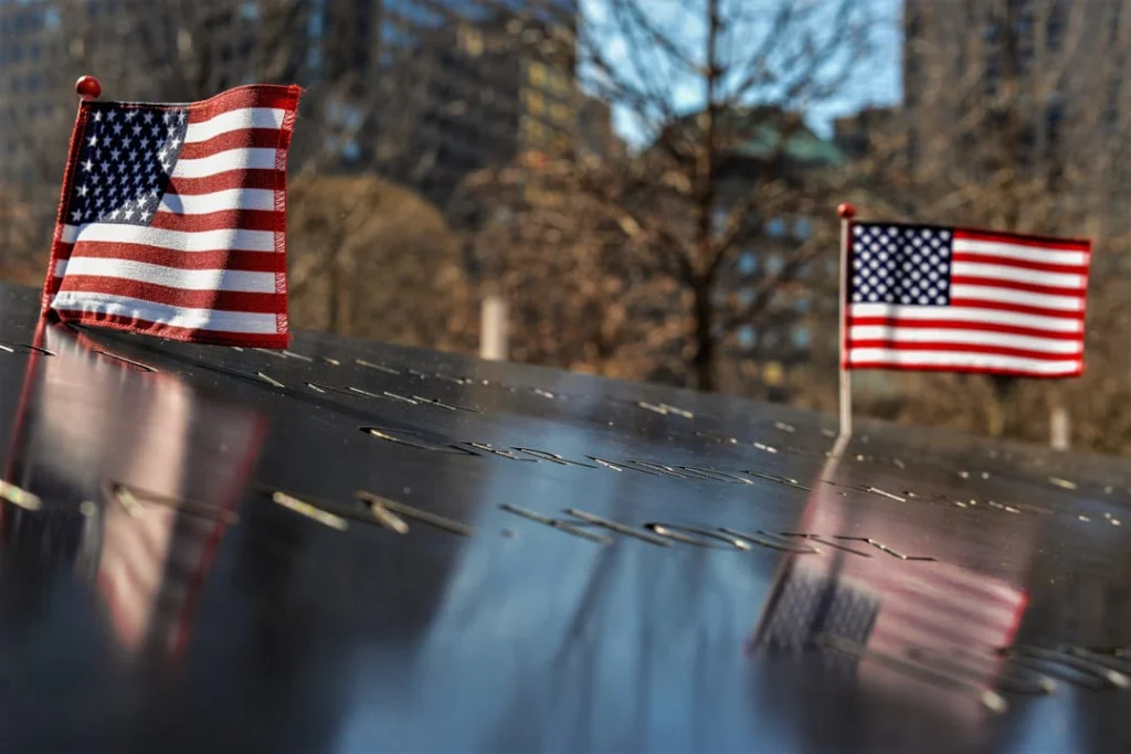 Kleine Amerikaanse vlaggetjes gestoken op de bronzen rand van het 9/11 Memorial in New York, met wazige gebouwen erachter