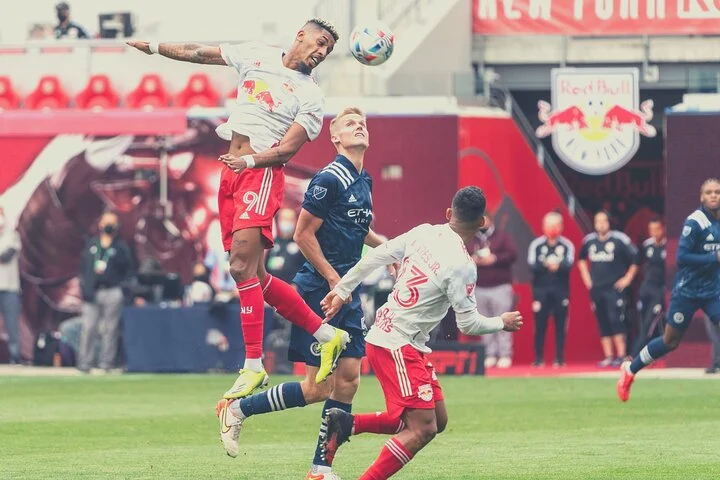 Voetballer springt op en kopt de bal terwijl tegenstanders proberen te reageren op het veld van de Red Bull Arena