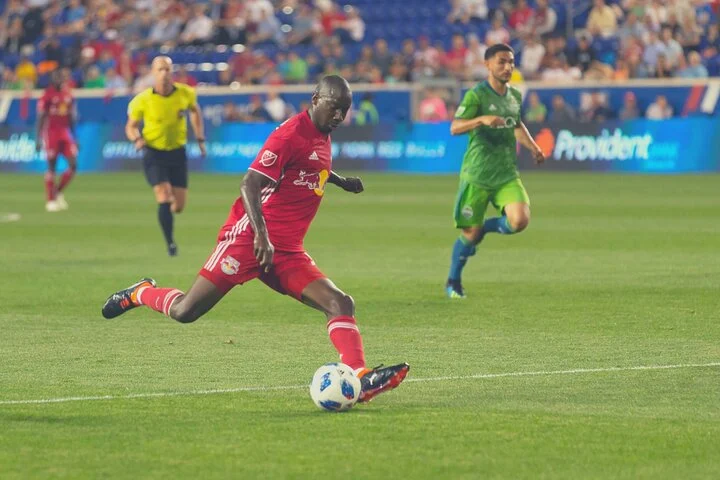 Voetballer in het rode shirt van New York Red Bulls haalt uit om op doel te schieten terwijl tegenstanders toekijken