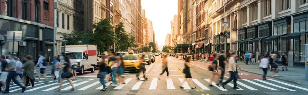 Drukke oversteekplaats op Fifth Avenue met voetgangers die de straat oversteken tussen hoge gebouwen terwijl de zon achter de straat ondergaat