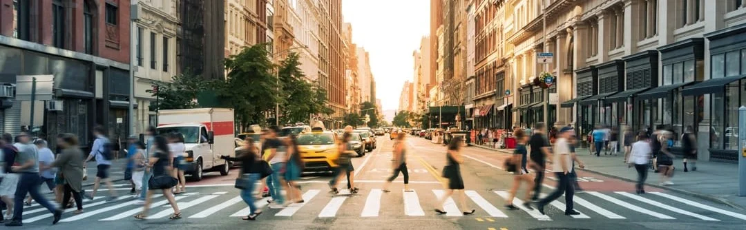 Drukke oversteekplaats op Fifth Avenue met voetgangers die de straat oversteken tussen hoge gebouwen terwijl de zon achter de straat ondergaat
