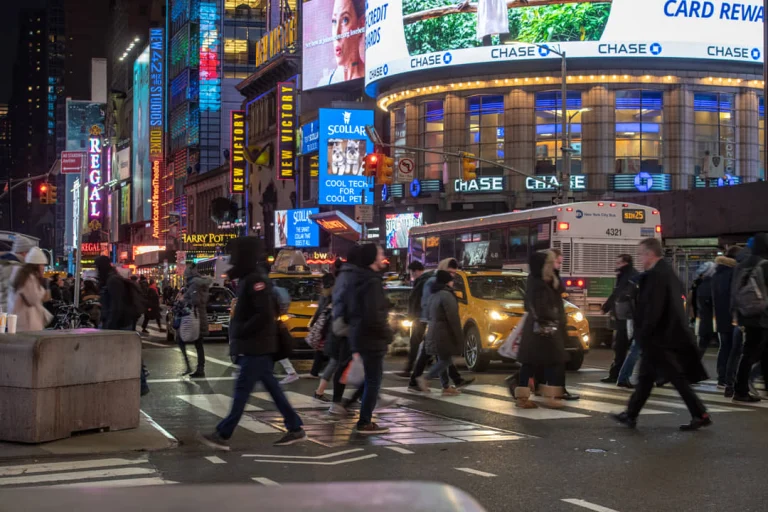 Grote groep voetgangers steekt een zebrapad over op Times Square met verlichte reclameborden en een bus bij schemering