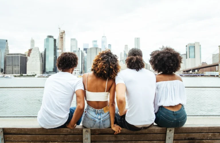 Vier vrienden zitten met hun rug naar de camera op een bankje aan het water en kijken uit over de wolkenkrabbers van de skyline van New York