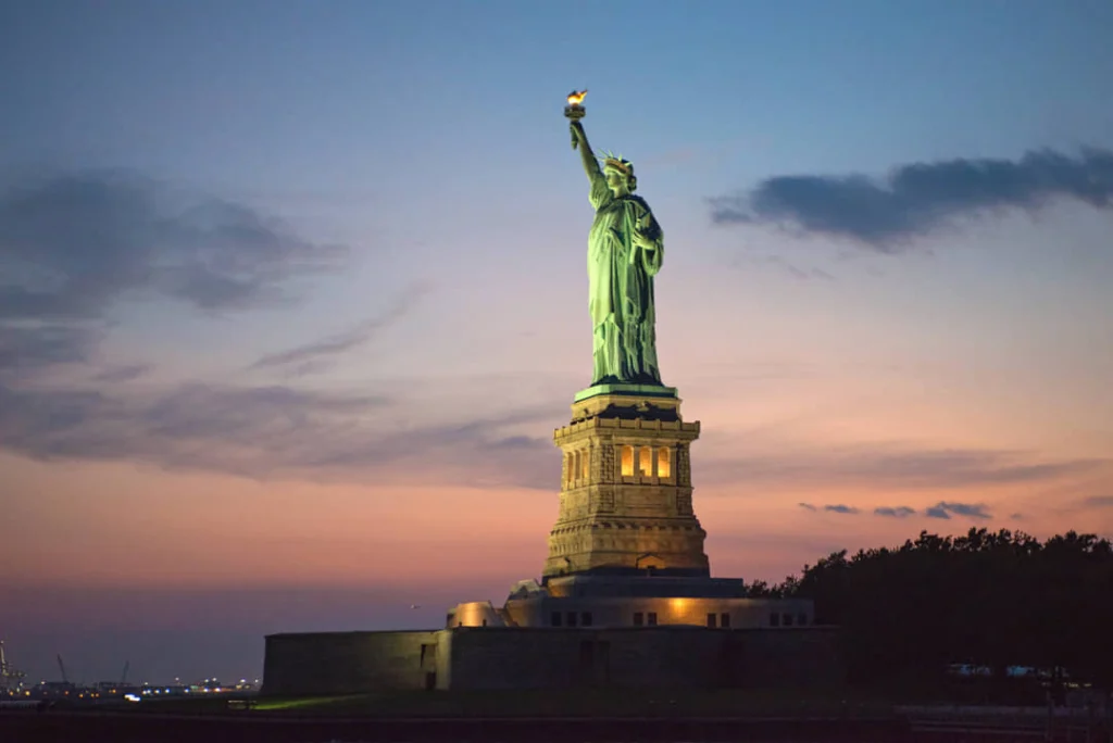 Het verlichte Vrijheidsbeeld in New York steekt af tegen een roze en paarse avondlucht bij zonsondergang