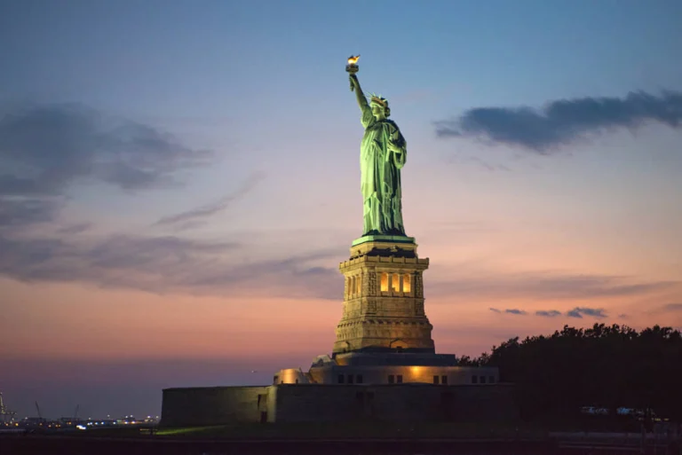 Het verlichte Vrijheidsbeeld in New York steekt af tegen een roze en paarse avondlucht bij zonsondergang