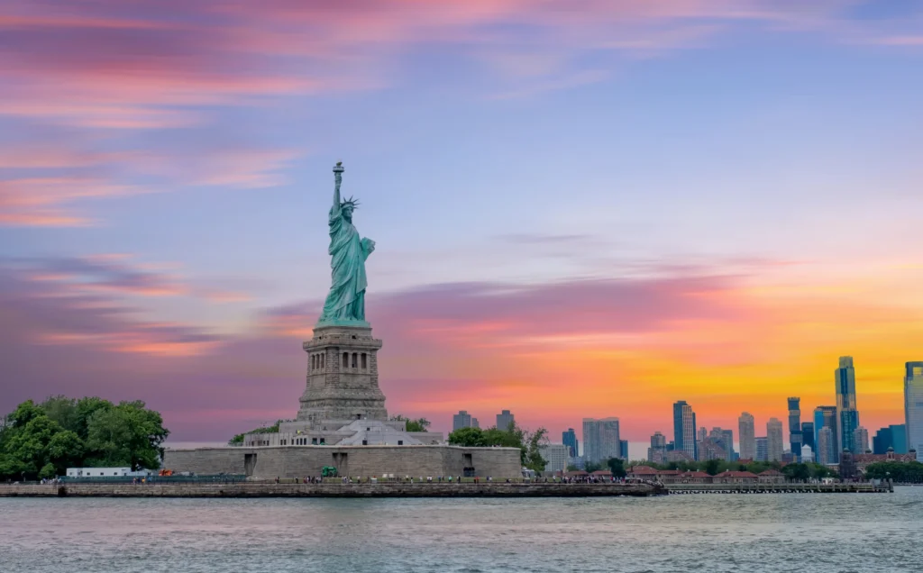 Het Vrijheidsbeeld op zijn voetstuk in de baai van New York met de skyline en een roze oranje avondlucht